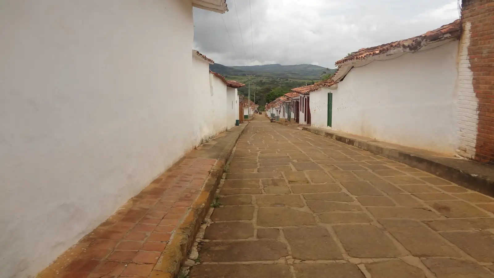 Calle de lajas de piedra, vacía, entre tapias blancas y aleros de teja, con andenes de ladrillo y montañas al fondo bajo un cielo nublado