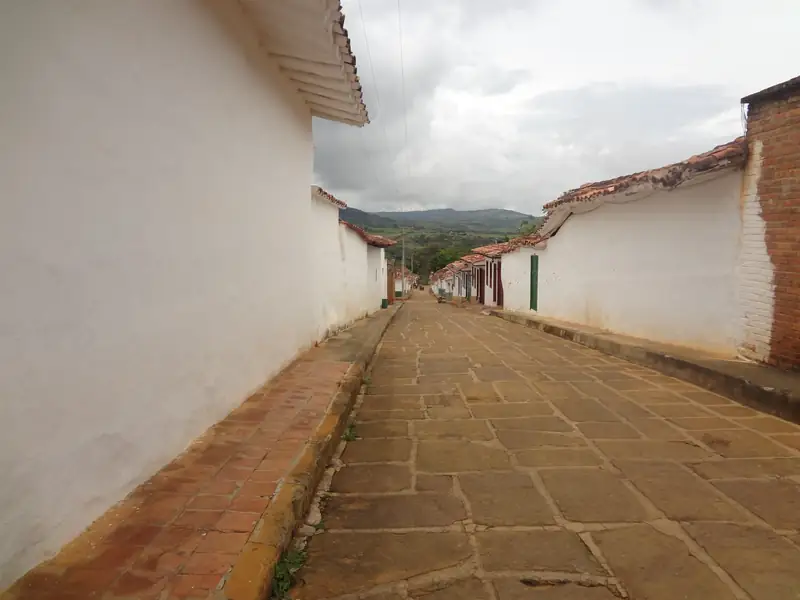 Calle de lajas de piedra, vacía, entre tapias blancas y aleros de teja, con andenes de ladrillo y montañas al fondo bajo un cielo nublado