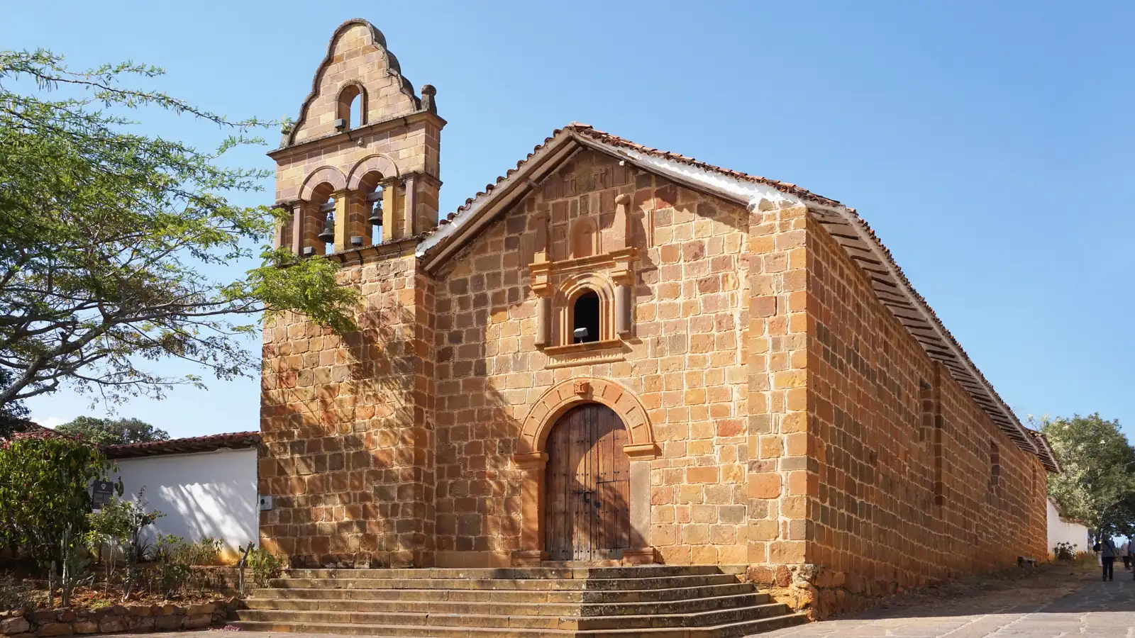 Capilla de piedra con espadaña de dos campanas, puerta de madera en arco y escalinata, bajo un cielo azul