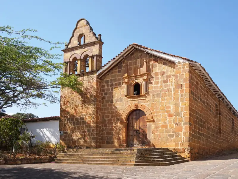Capilla de piedra con espadaña de dos campanas, puerta de madera en arco y escalinata, bajo un cielo azul