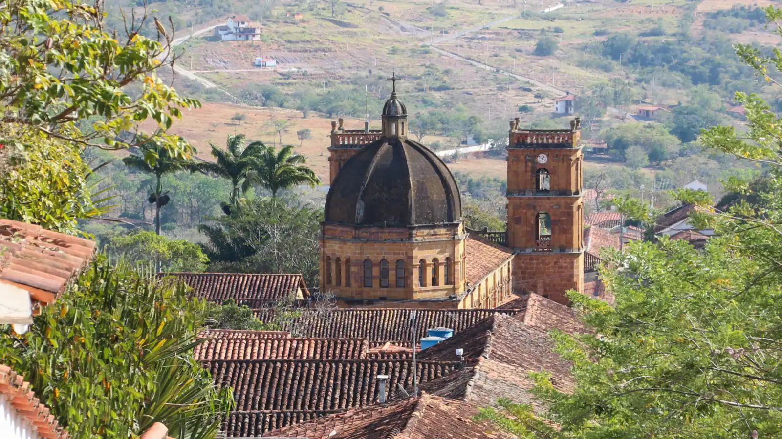 La cúpula y las torres del templo asoman sobre techos de teja de barro, entre árboles, con la ladera del valle al fondo
