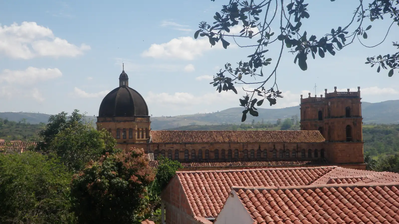 Cúpula y torres del templo de piedra de Barichara sobre los techos de teja, con montañas verdes al fondo y una rama en primer plano