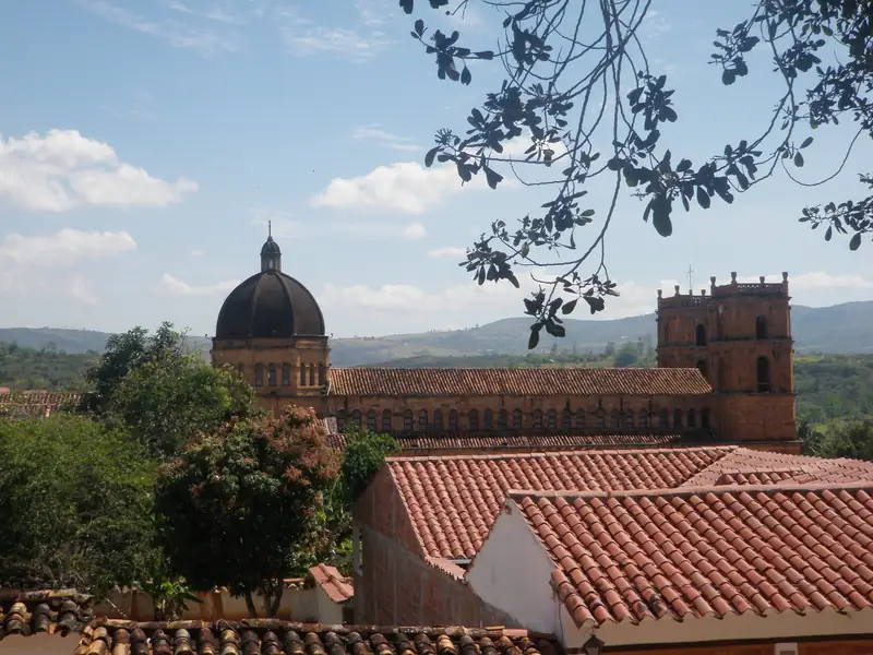 Cúpula y torres del templo de piedra de Barichara sobre los techos de teja, con montañas verdes al fondo y una rama en primer plano