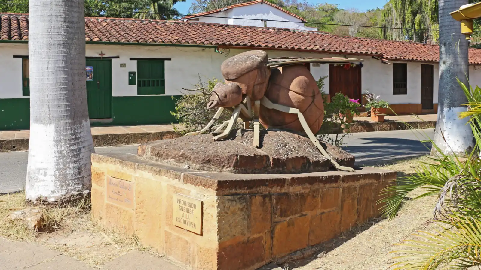 Escultura de una hormiga culona sobre una base de piedra, con patas y alas de metal, frente a casas blancas de puertas verdes