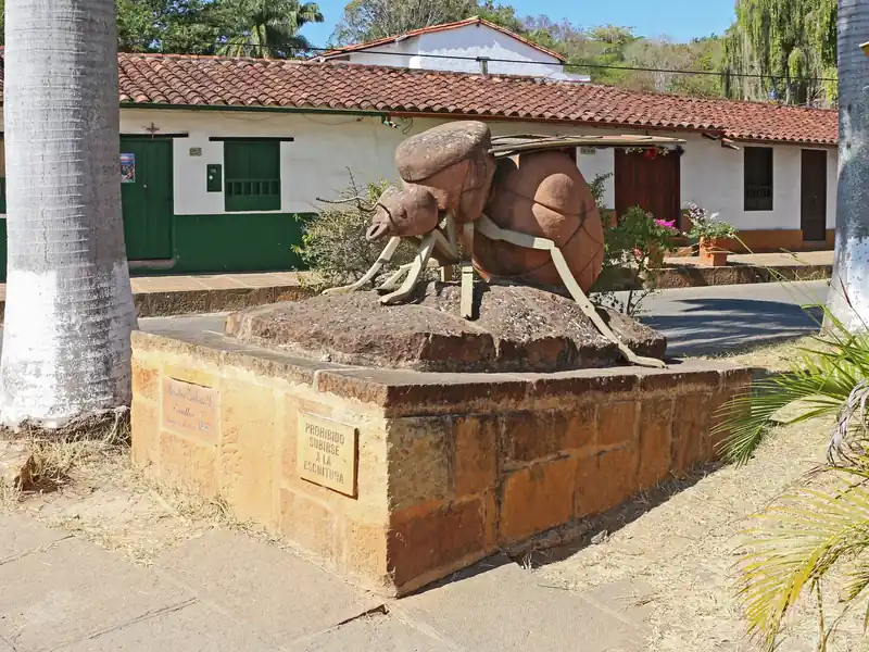 Escultura de una hormiga culona sobre una base de piedra, con patas y alas de metal, frente a casas blancas de puertas verdes