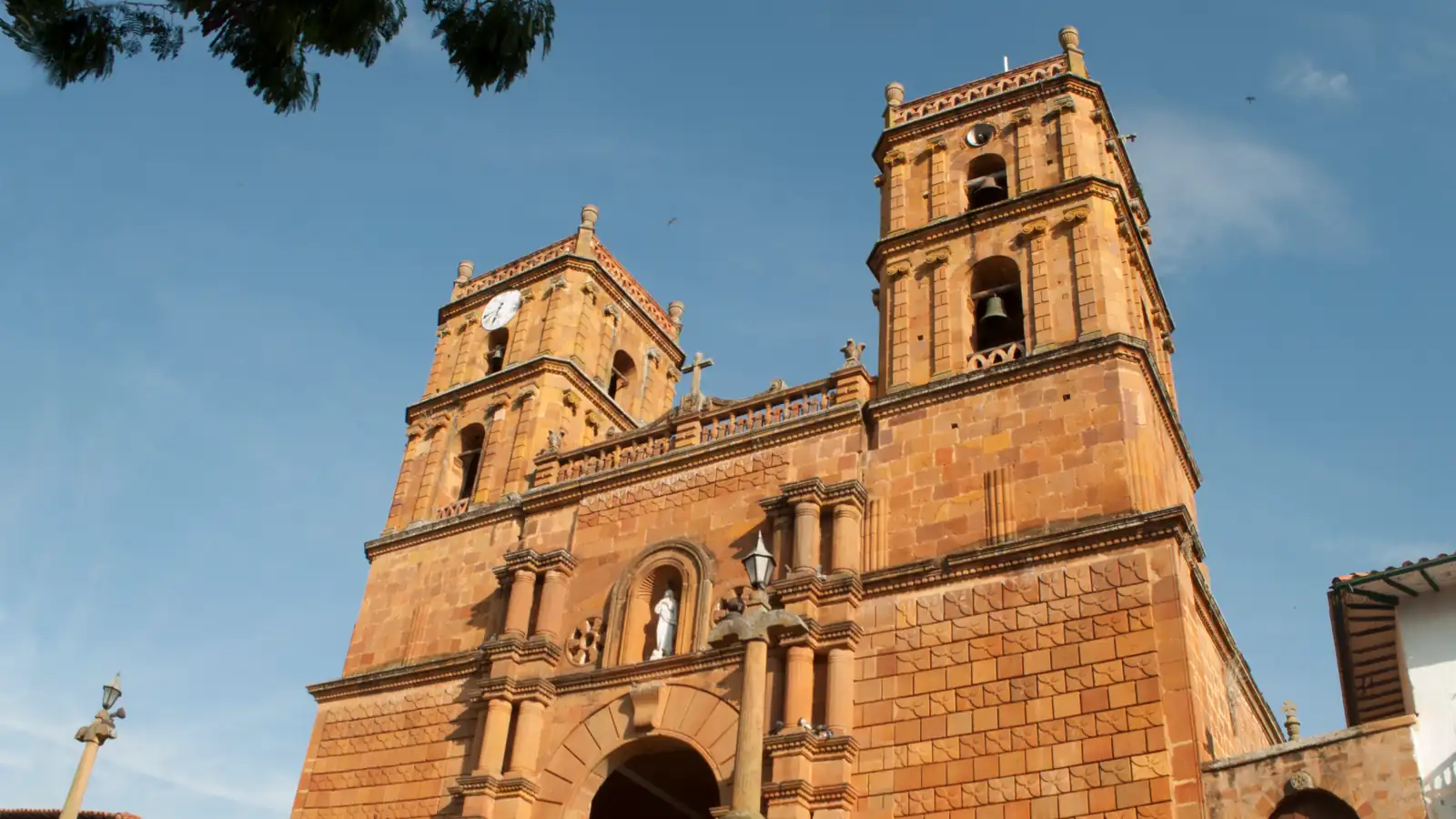Fachada de piedra labrada del templo, con sus dos torres de campanario, una imagen blanca en el nicho sobre la puerta y la escalinata delante