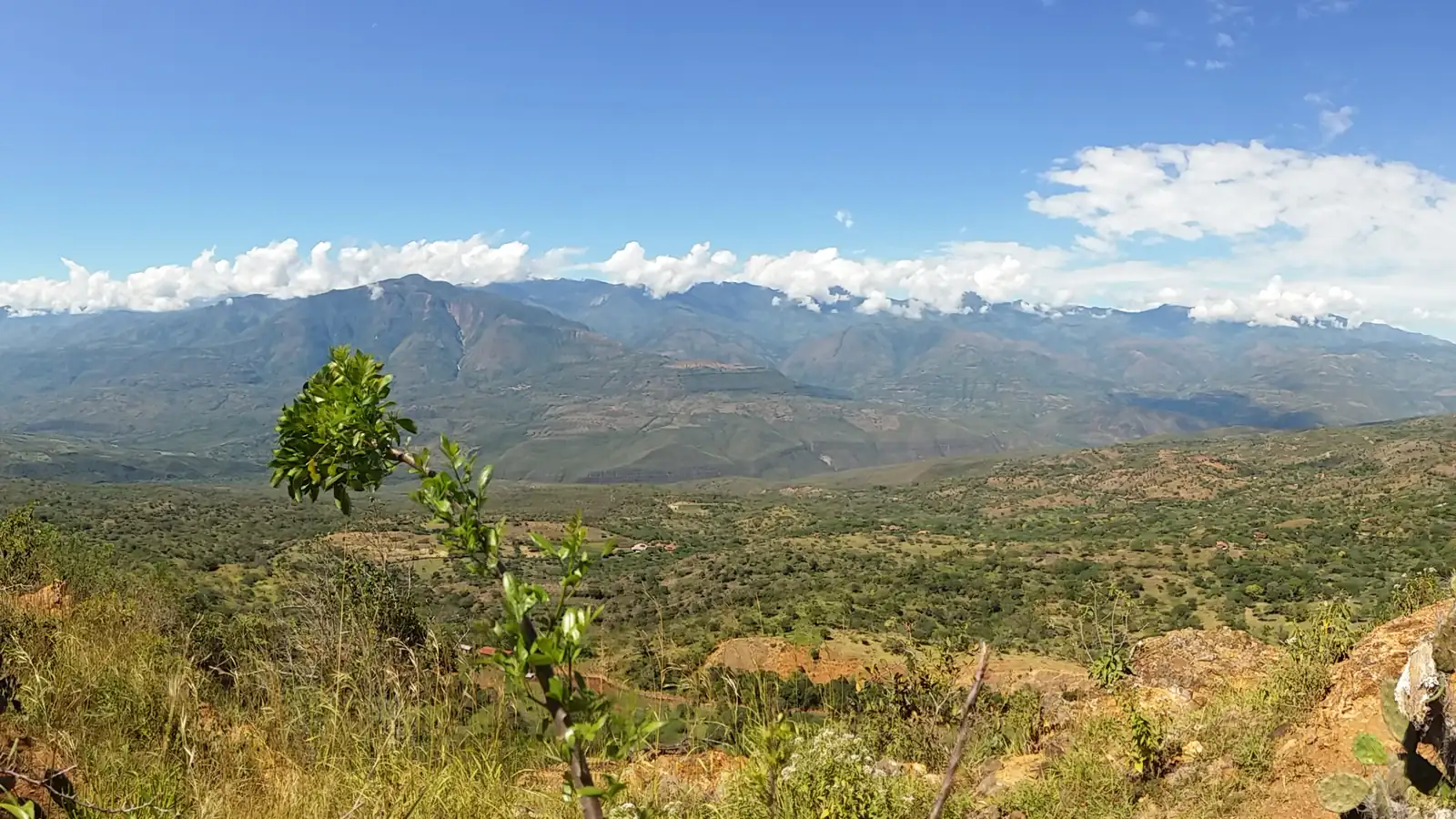 Vista desde el borde occidental de Barichara: el valle con monte seco abajo y, al fondo, una cordillera con nubes blancas bajo el cielo azul