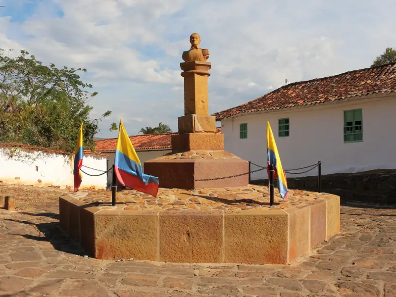 Busto de Simón Bolívar sobre un pedestal de piedra amarilla, con tres banderas de Colombia, en una plazoleta empedrada entre casas blancas