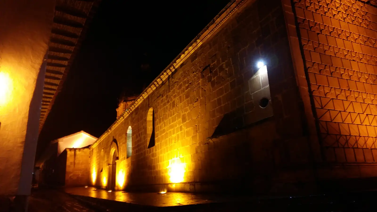 Muro lateral de piedra del templo de la Inmaculada Concepción, iluminado de noche desde abajo, junto a una calle empedrada y el alero de una casa