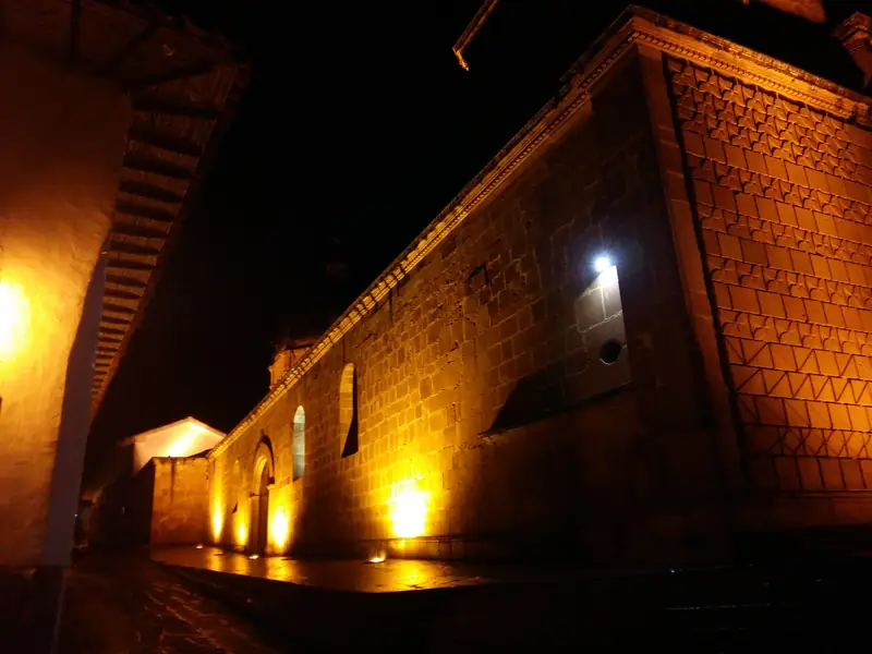 Muro lateral de piedra del templo de la Inmaculada Concepción, iluminado de noche desde abajo, junto a una calle empedrada y el alero de una casa