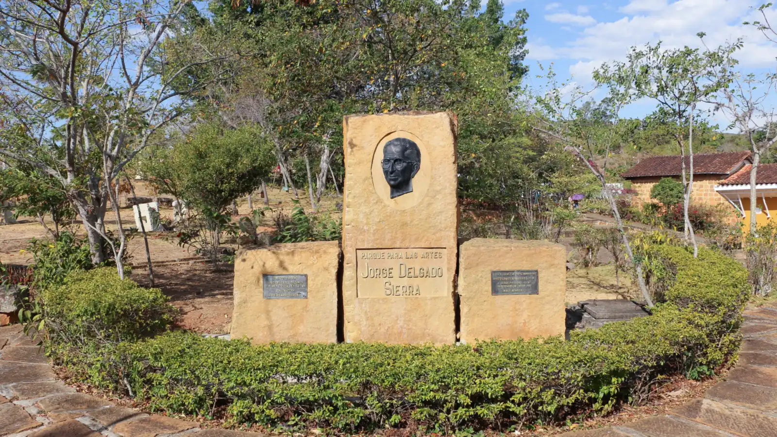 Monumento de bloques de piedra con un retrato en relieve y la inscripción «Parque para las Artes Jorge Delgado Sierra», rodeado de setos, árboles y un andén de lajas