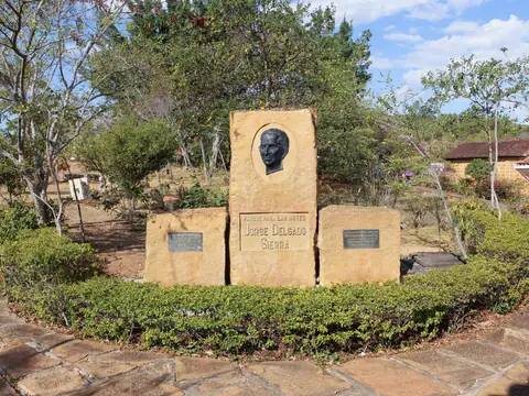 Monumento de bloques de piedra con un retrato en relieve y la inscripción «Parque para las Artes Jorge Delgado Sierra», rodeado de setos, árboles y un andén de lajas