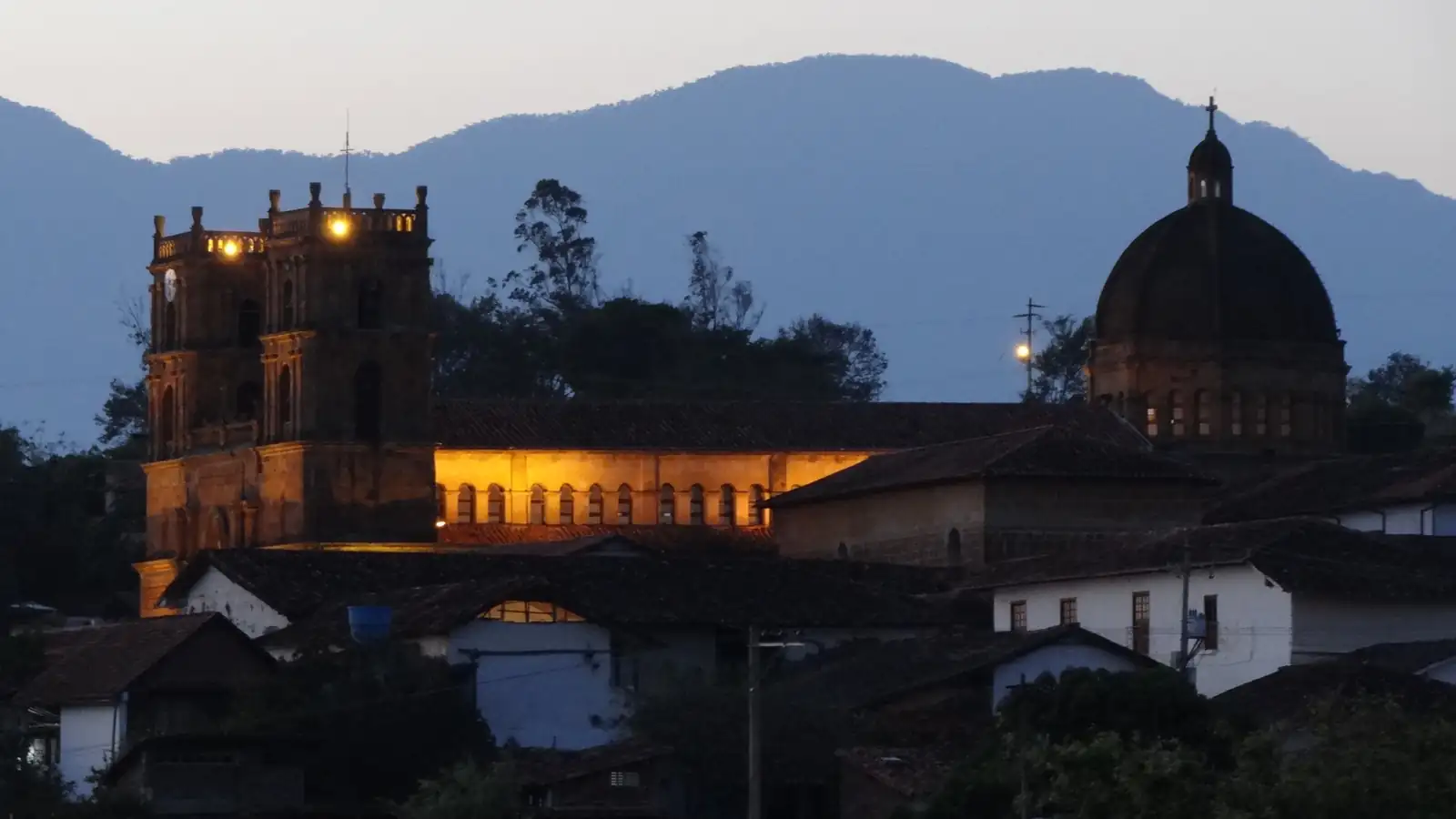 El templo de la Inmaculada Concepción iluminado al anochecer, con sus dos torres, la nave y la cúpula sobre los techos de teja y las montañas azules detrás