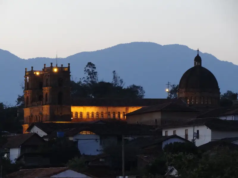 El templo de la Inmaculada Concepción iluminado al anochecer, con sus dos torres, la nave y la cúpula sobre los techos de teja y las montañas azules detrás