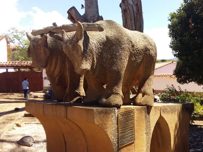 Escultura de una yunta de bueyes tallada en piedra sobre un pedestal de piedra con una placa de bronce, entre árboles y casas de techo de teja