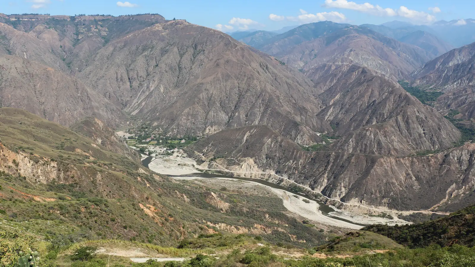 El río Chicamocha serpentea entre montañas áridas y pardas del cañón, bajo un cielo azul con pocas nubes y con matorral verde en primer plano