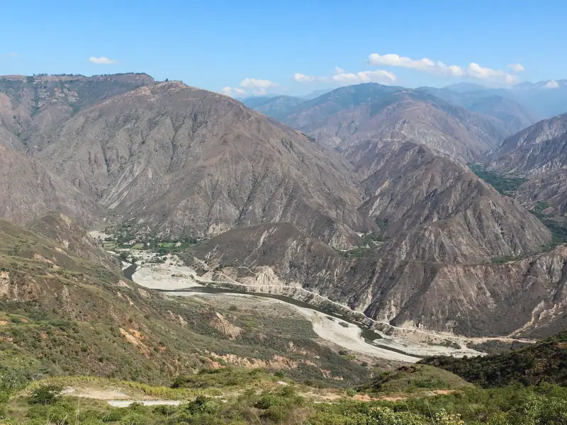El río Chicamocha serpentea entre montañas áridas y pardas del cañón, bajo un cielo azul con pocas nubes y con matorral verde en primer plano