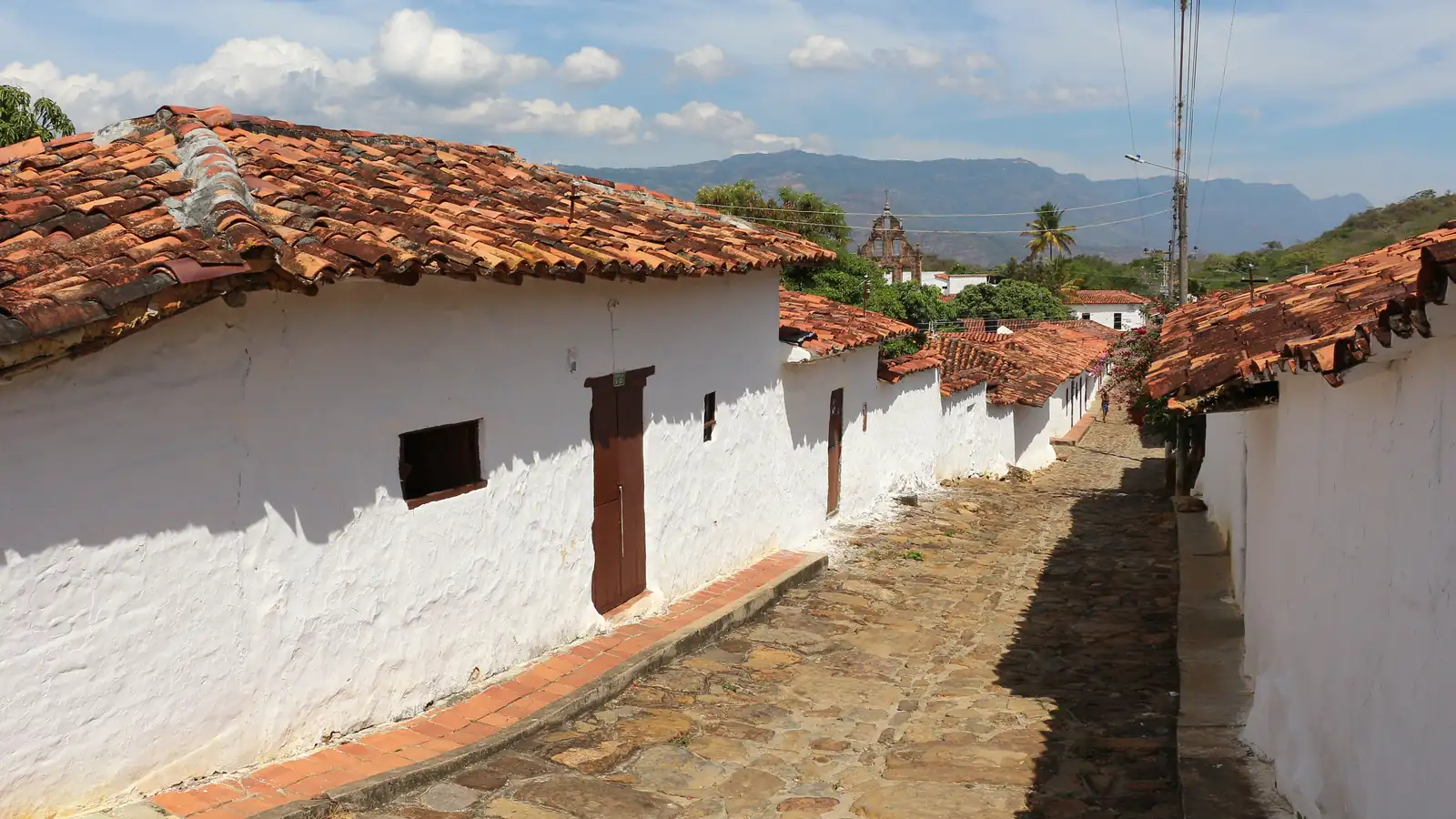 Calle empedrada de Guane entre casas blancas con techos de teja de barro; al fondo, la espadaña de la iglesia y las montañas