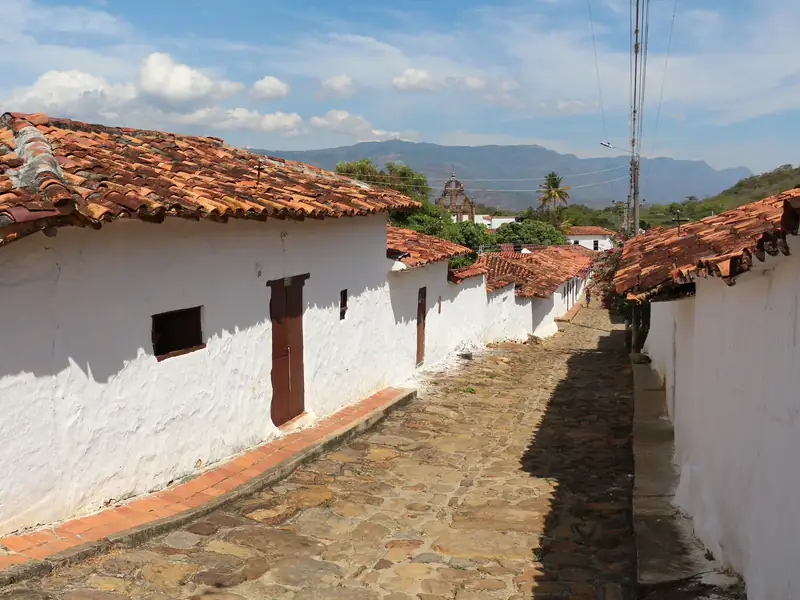 Calle empedrada de Guane entre casas blancas con techos de teja de barro; al fondo, la espadaña de la iglesia y las montañas