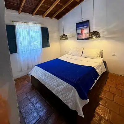 Cama queen con cobija azul bajo techo de madera y ventana azul, en la Habitación Doble