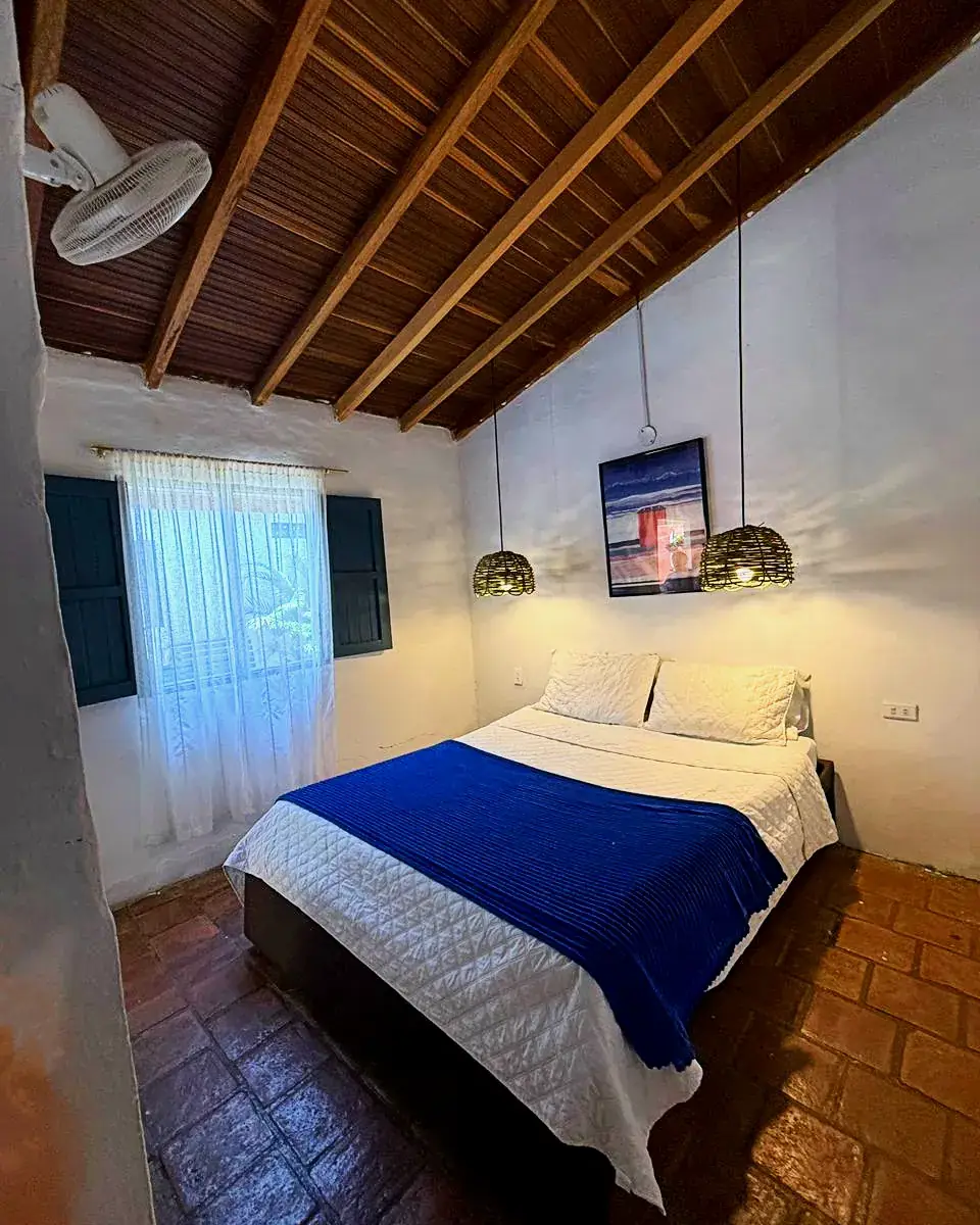 Cama queen con cobija azul bajo techo de madera y ventana azul, en la Habitación Doble
