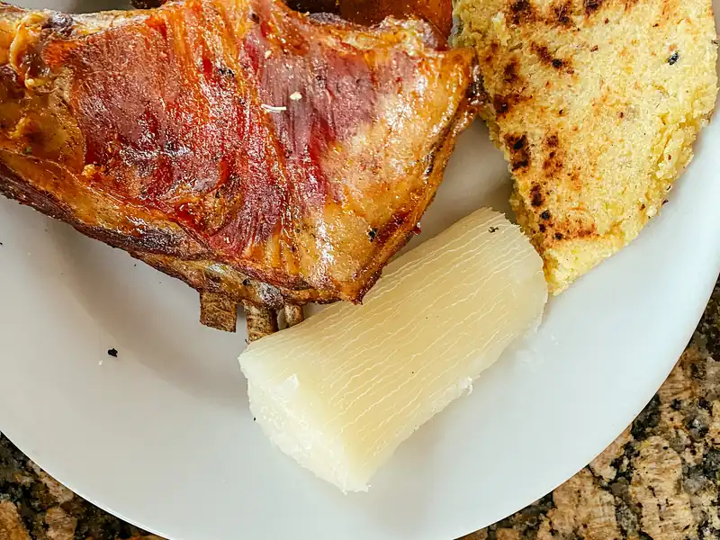 Cabro al horno con un trozo de yuca y una arepa tostada en un plato blanco; al lado, otro plato con pepitoria de arroz