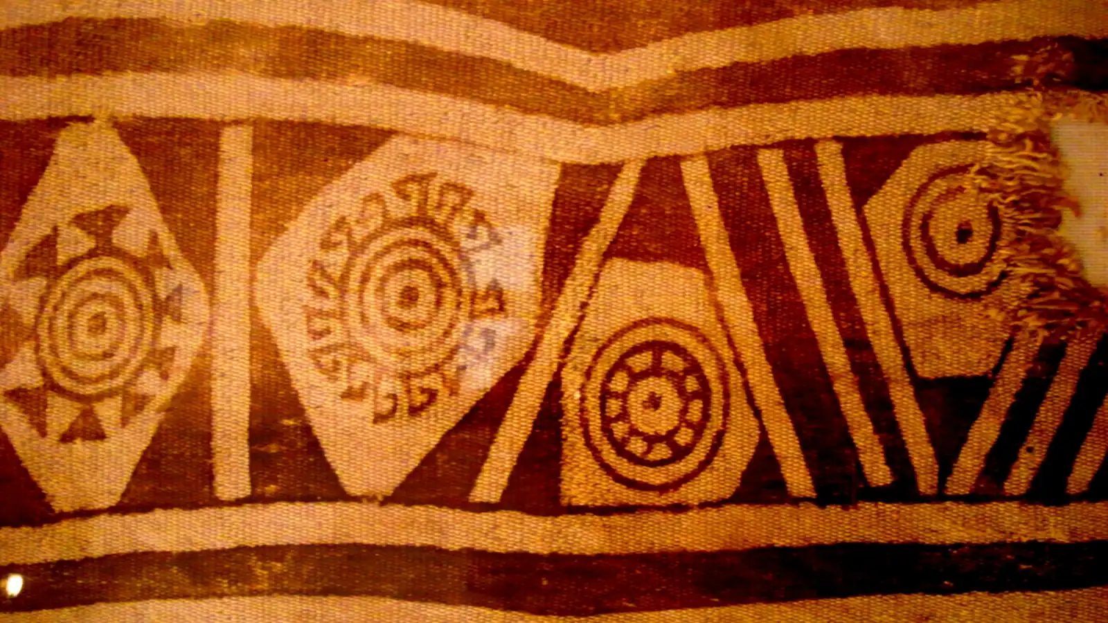 Fragmento de tela antigua con rombos, círculos concéntricos y franjas en café y crema, descrito en Wikimedia Commons como un tejido guane