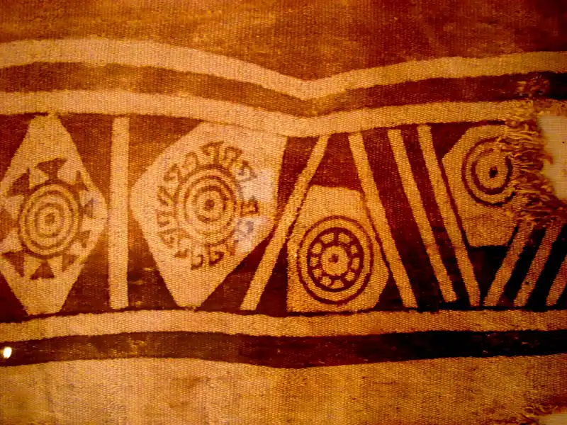 Fragmento de tela antigua con rombos, círculos concéntricos y franjas en café y crema, descrito en Wikimedia Commons como un tejido guane