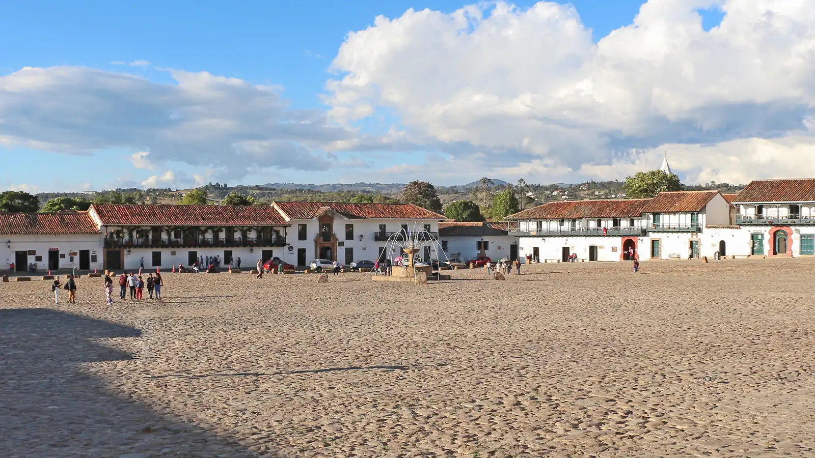 La Plaza Mayor de Villa de Leyva, empedrada y abierta, con casas blancas de balcones y teja alrededor y una fuente de piedra en el centro, bajo nubes grandes