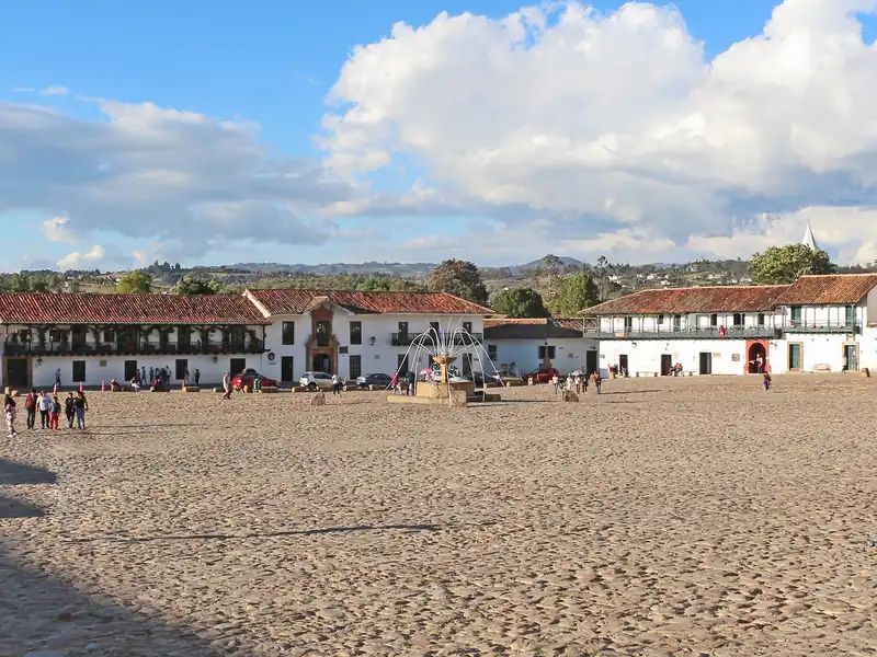 La Plaza Mayor de Villa de Leyva, empedrada y abierta, con casas blancas de balcones y teja alrededor y una fuente de piedra en el centro, bajo nubes grandes