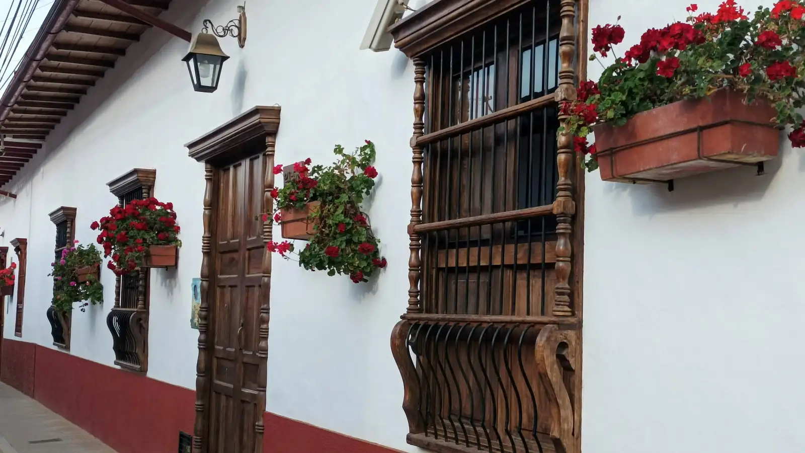 Fachada blanca de Zapatoca con zócalo rojo, puerta y ventanas de madera tallada y materas de geranios rojos sobre la acera