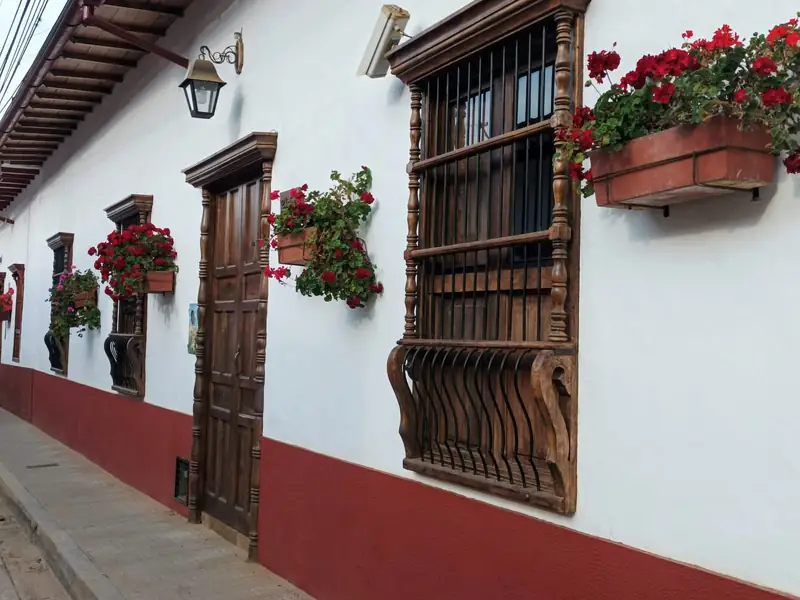 Fachada blanca de Zapatoca con zócalo rojo, puerta y ventanas de madera tallada y materas de geranios rojos sobre la acera